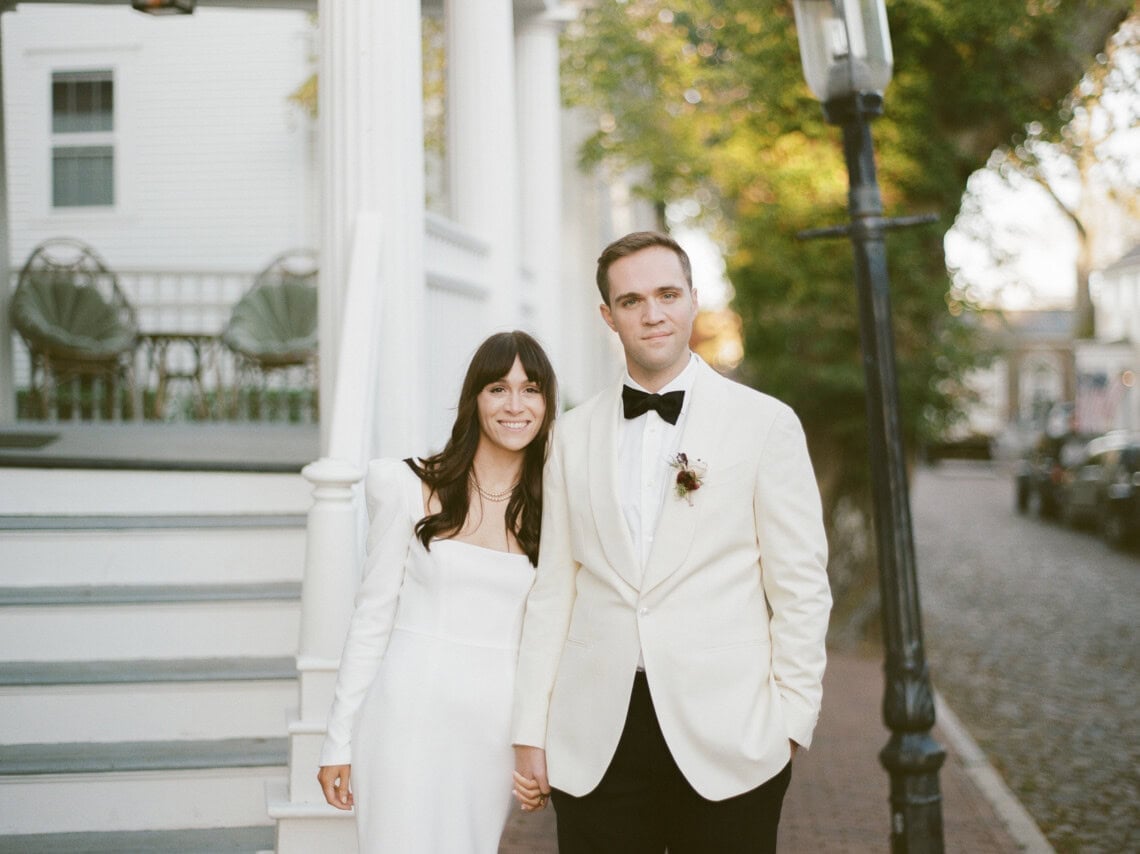 A bride in a white dress and a groom in a white tuxedo jacket stand hand-in-hand on a brick sidewalk near white steps, smiling at the camera, beautifully captured by a Nantucket photographer, with trees and a street in the background.