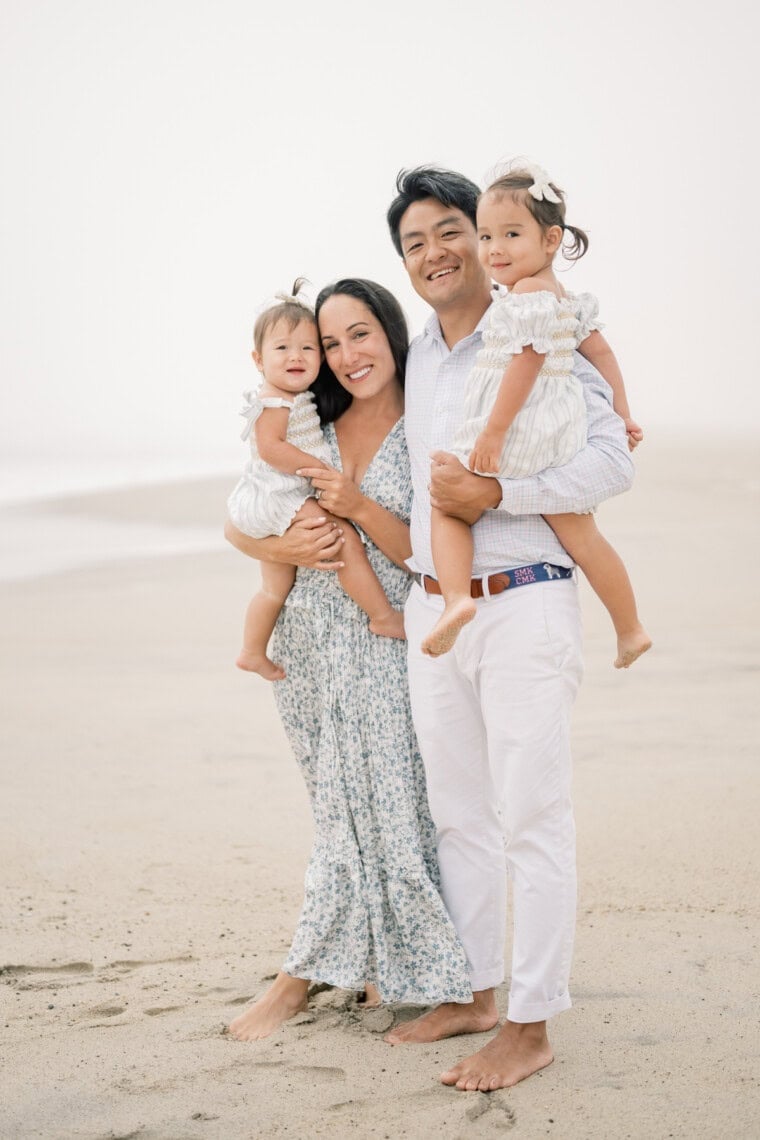 A smiling family of four stands barefoot on a sandy beach, captured by a Nantucket photographer. The mother holds a young toddler, and the father an older girl. Both children wear white dresses and hair bows; all are dressed in light, summery clothing.