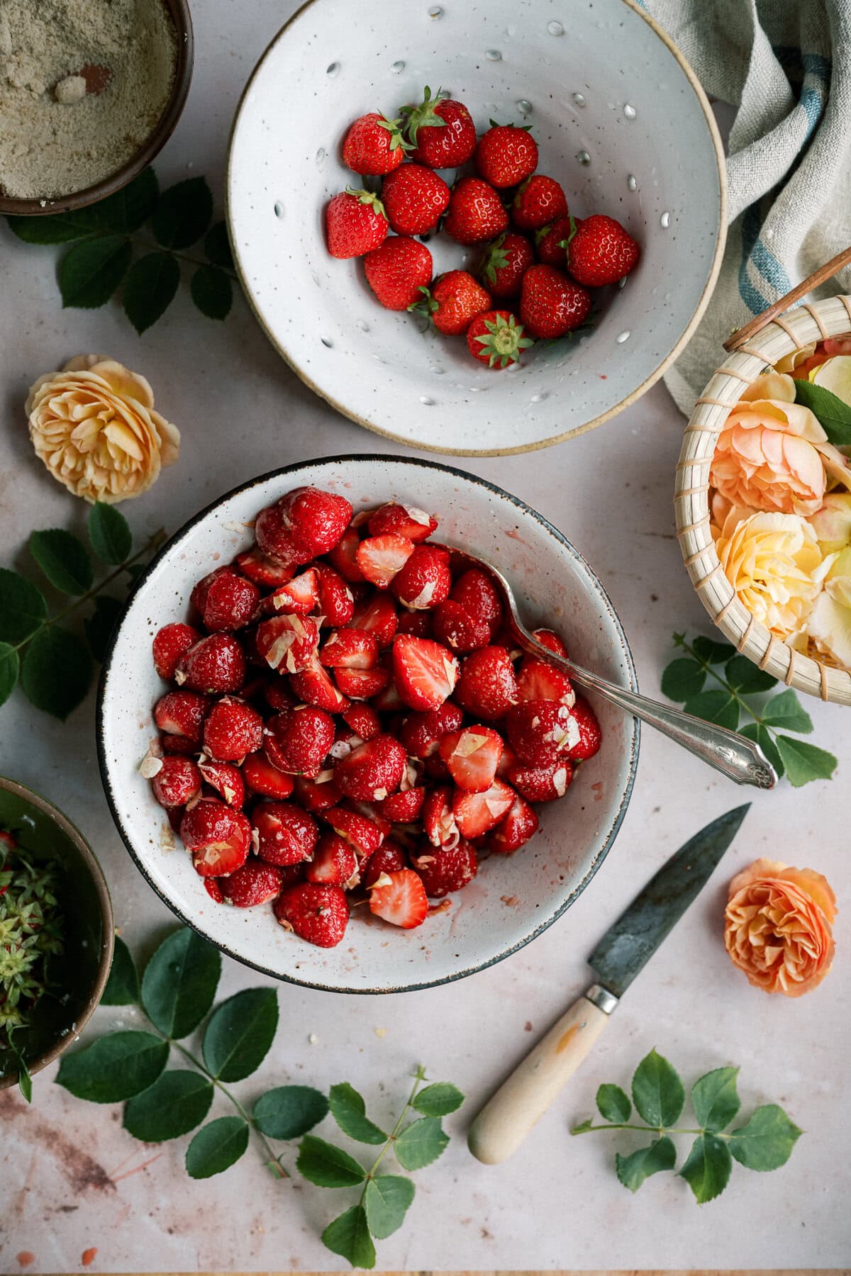 A bowl of sliced strawberries with a spoon sits on a table surrounded by whole strawberries, yellow roses, green leaves, a knife, and a basket with more flowers—perfect for making strawberry shortcake with biscuits.