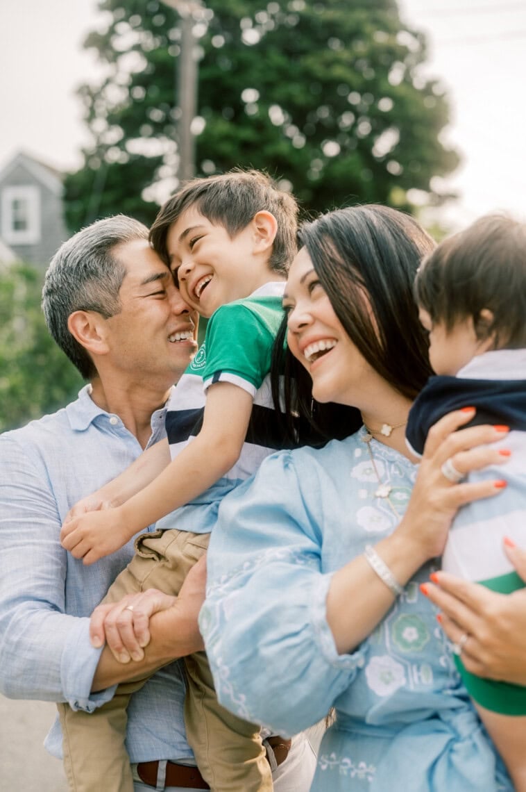 A joyful family of four smiles and laughs together outdoors as their moments are beautifully captured by a Nantucket photographer. The father holds a young boy, while the mother hugs a smaller child. Trees and a house are visible in the background.