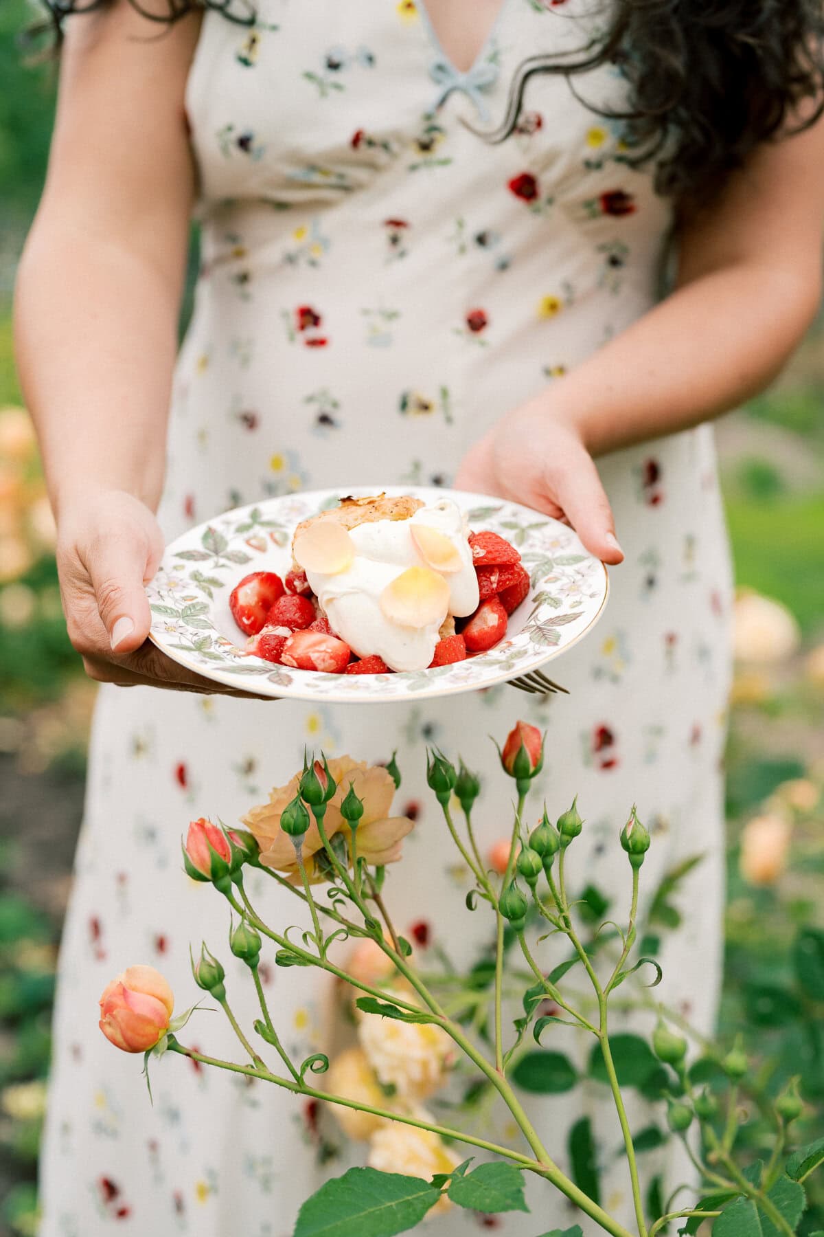 A woman in a floral dress holds a plate with strawberry shortcake with biscuits, topped with cream and edible flower petals, standing in a garden filled with blooming roses.