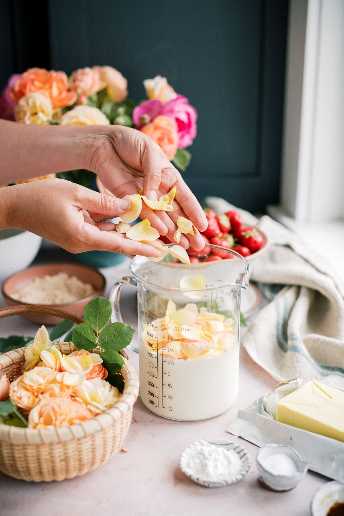 Hands sprinkle yellow rose petals into a glass measuring cup filled with milk. Nearby are fresh flowers, butter, flour, sugar, and a bowl of strawberries—ingredients ready for homemade strawberry shortcake with biscuits.