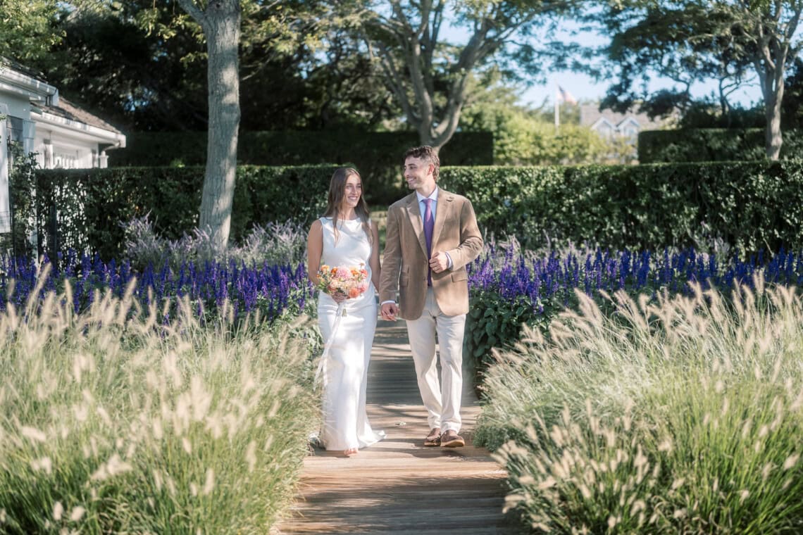 A bride in a white dress holding a bouquet and a groom in a tan blazer walk together on a wooden path, captured by a Nantucket photographer, surrounded by tall grass and purple flowers under a sunny, blue sky.