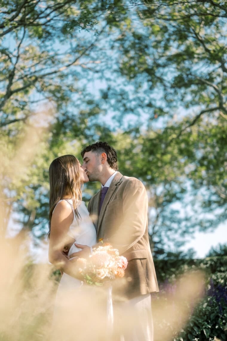 A couple dressed in wedding attire shares a gentle kiss outdoors, surrounded by greenery and sunlight, with the bride holding a bouquet of flowers—captured beautifully by a Nantucket photographer.