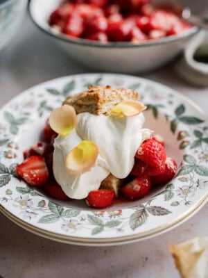 A dessert plate of strawberry shortcake with biscuits features sliced strawberries, fluffy whipped cream, and delicate flower petals on a floral-patterned dish. A bowl of strawberries is visible in the background.
