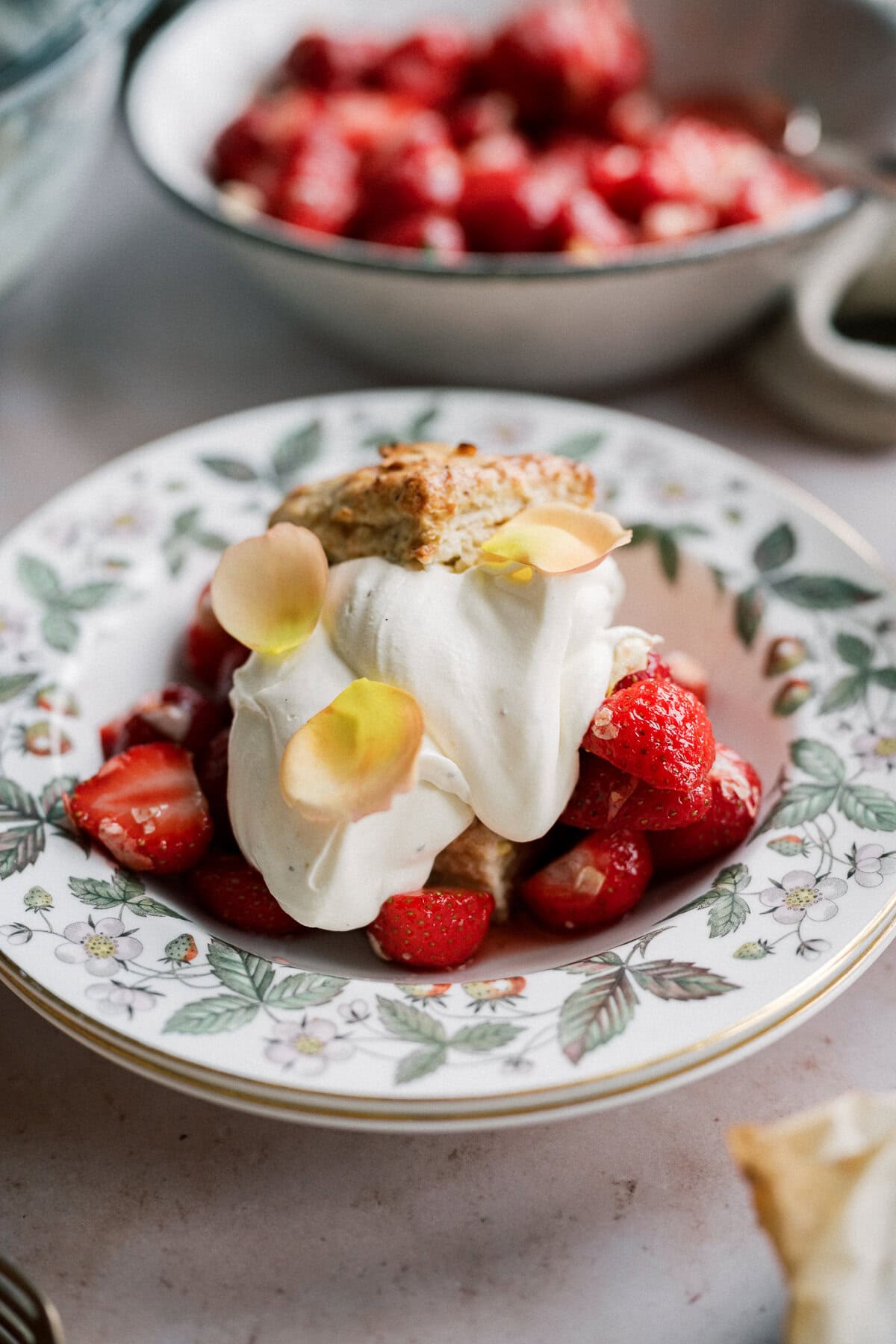 A dessert plate of strawberry shortcake with biscuits features sliced strawberries, fluffy whipped cream, and delicate flower petals on a floral-patterned dish. A bowl of strawberries is visible in the background.