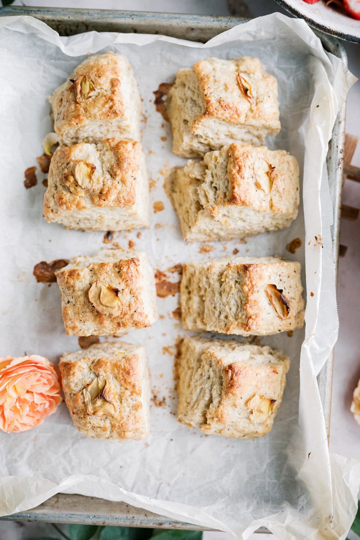 Six golden-brown, square biscuits—perfect for a strawberry shortcake with biscuits—are arranged on a parchment-lined tray. Crumbs are scattered around, and a peach-colored flower sits at the lower left corner.