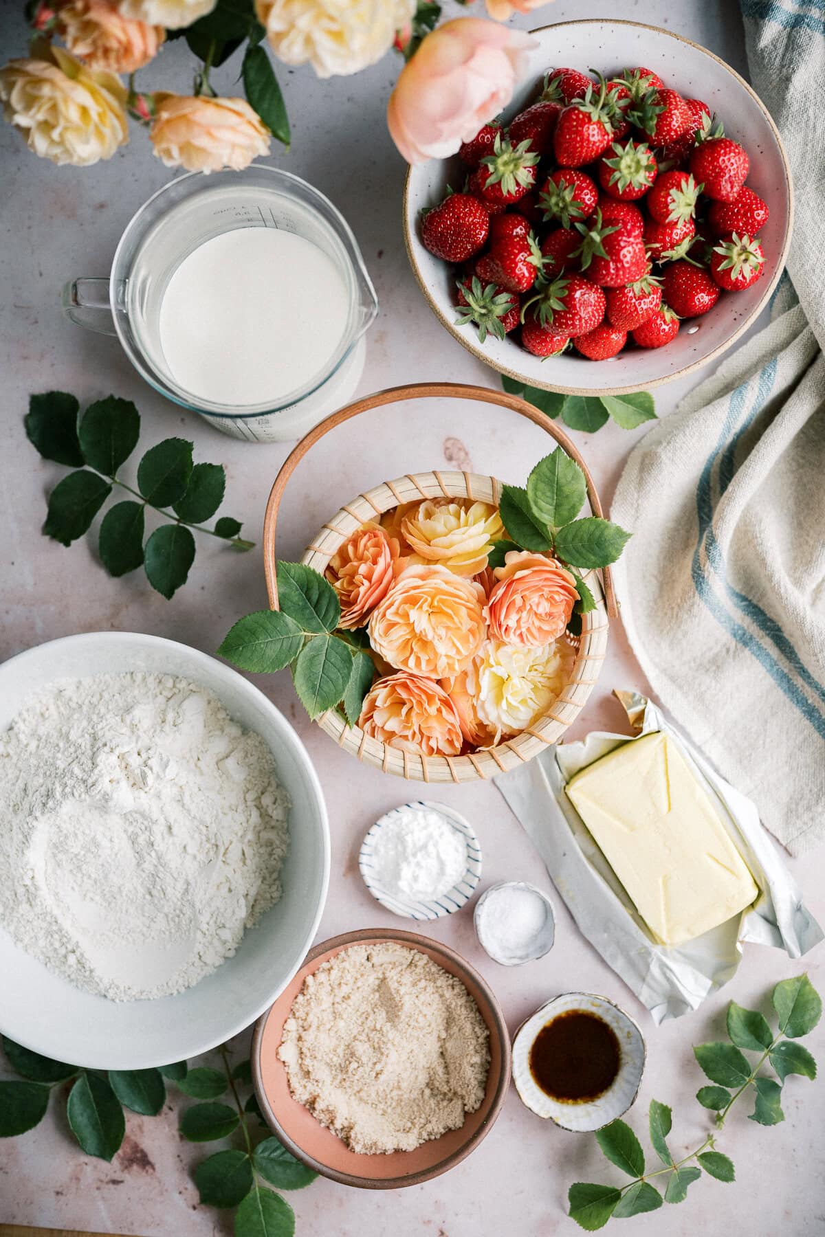 A flat lay of baking ingredients for strawberry shortcake with biscuits, including flour, brown sugar, butter, strawberries, vanilla, baking powder, salt, and milk, accented by yellow and peach roses with green leaves on a light surface.