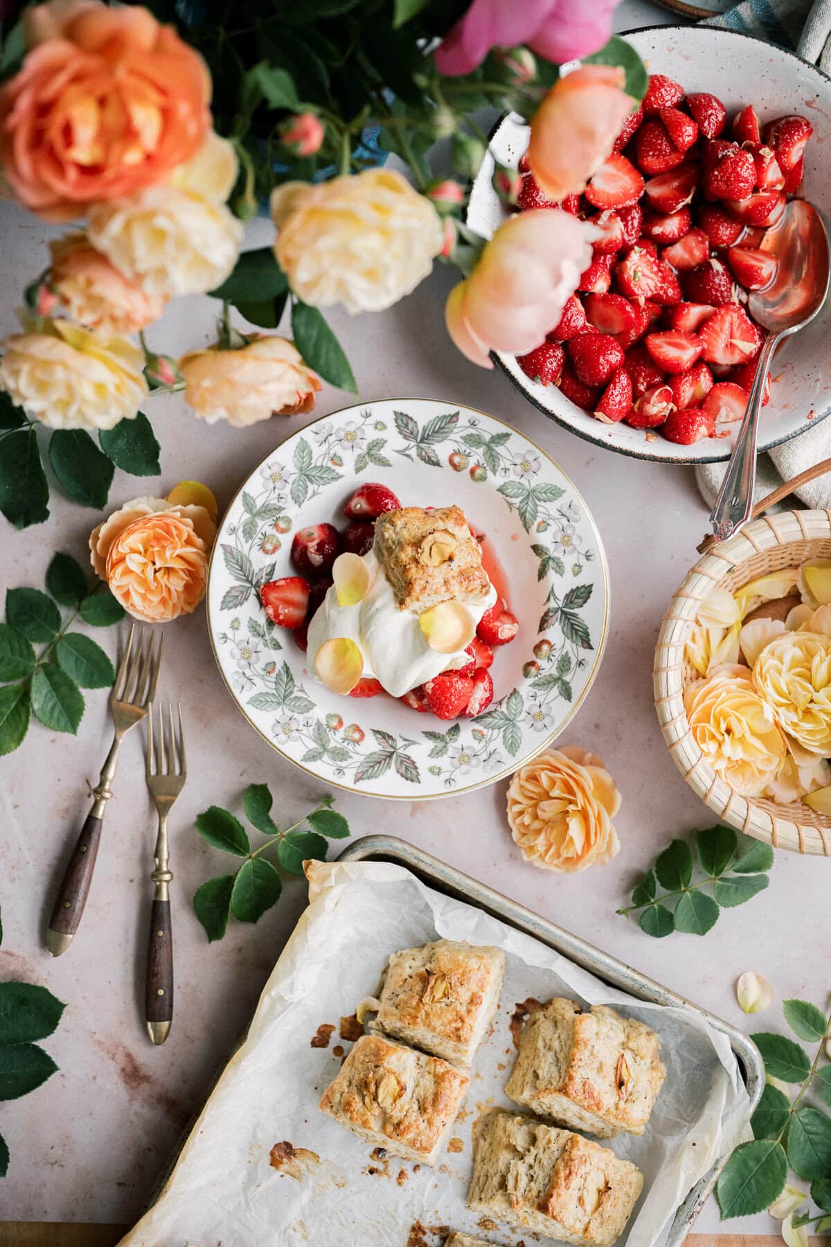 A floral table setting features strawberry shortcake with biscuits, topped with whipped cream and strawberries, surrounded by fresh strawberries, yellow roses, forks, and a tray of baked shortcakes on parchment paper.