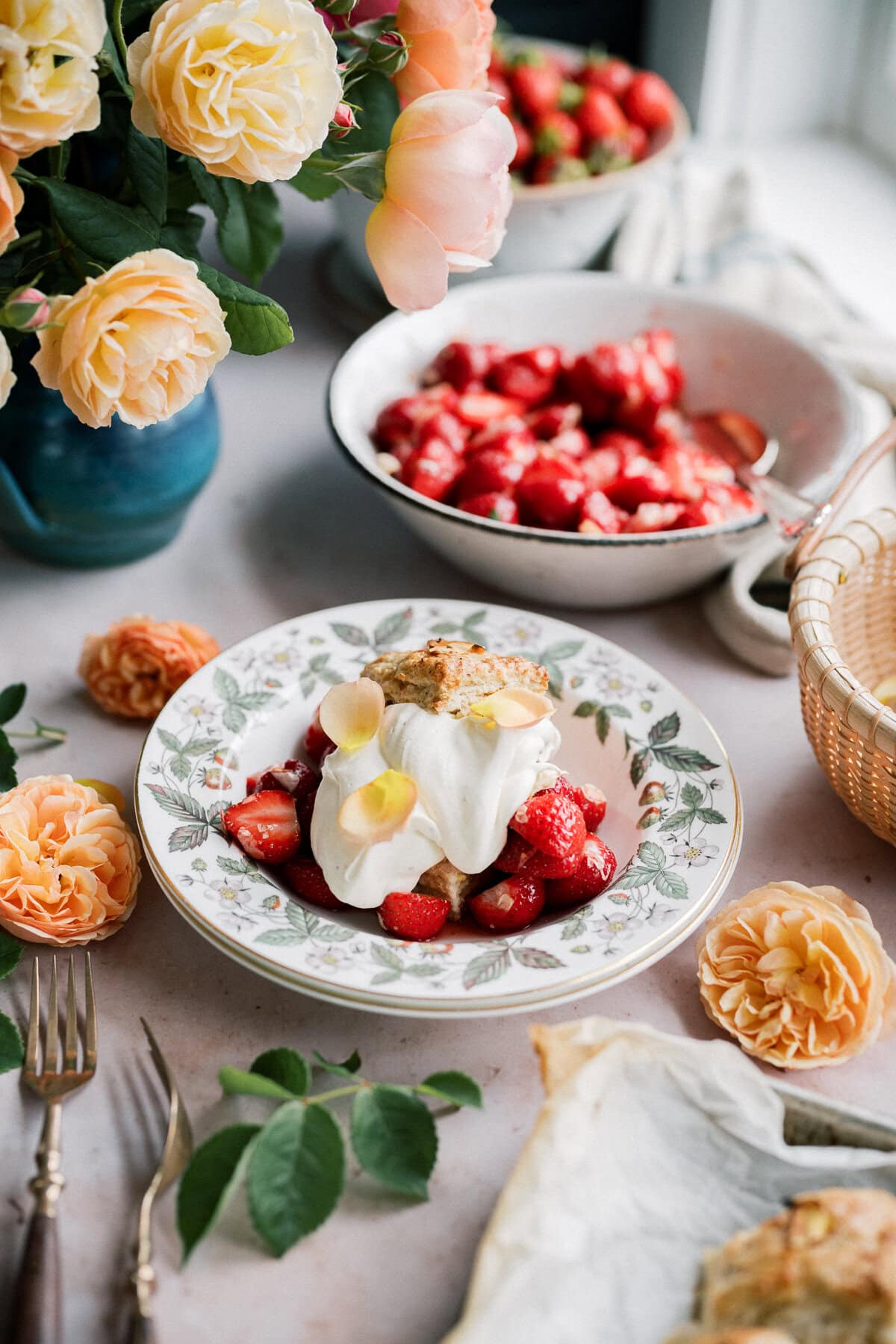 A bowl of strawberry shortcake with biscuits, topped with whipped cream and flower petals, sits on a floral-patterned plate. Yellow roses and more strawberries decorate the table, creating a bright, fresh, and elegant setting.