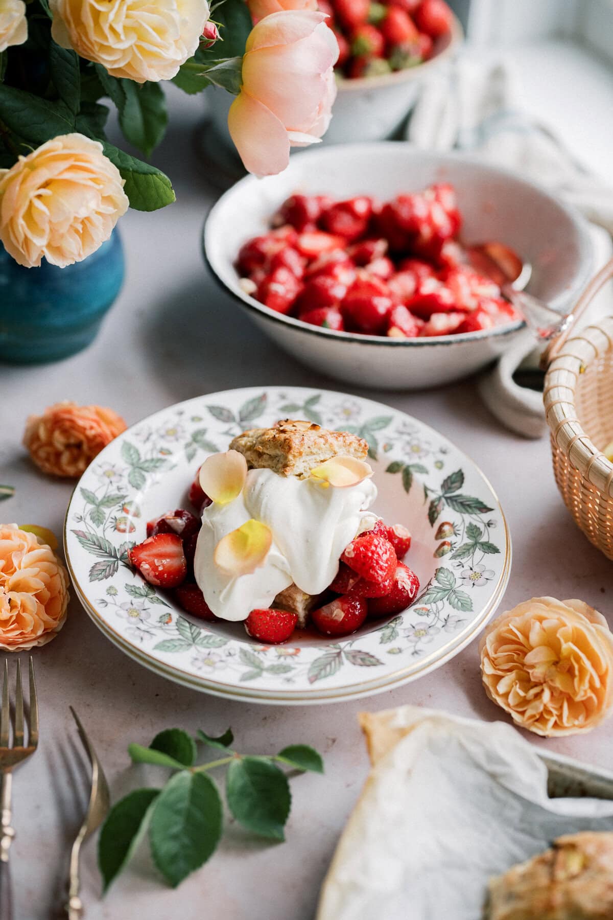 A bowl of strawberry shortcake with biscuits is topped with whipped cream and garnished with yellow flower petals, sitting on a floral plate. Fresh yellow roses and bowls of strawberries decorate the table.