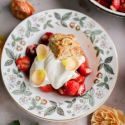 A strawberry shortcake with biscuits is served on a floral-patterned plate, topped with fresh strawberries, whipped cream, almond slices, and flower petals, surrounded by blooms for a charming dessert presentation.