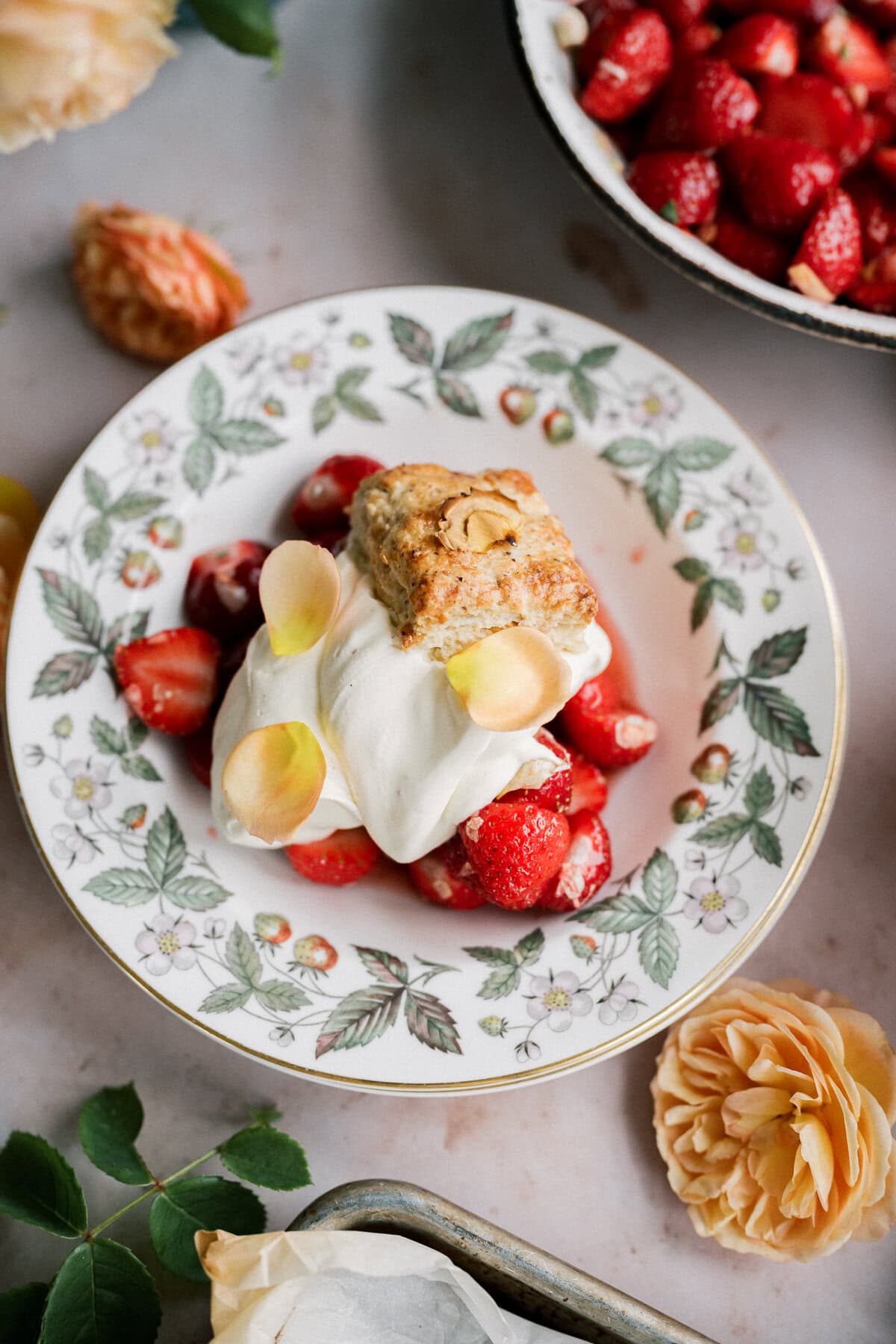 A strawberry shortcake with biscuits is served on a floral-patterned plate, topped with fresh strawberries, whipped cream, almond slices, and flower petals, surrounded by blooms for a charming dessert presentation.