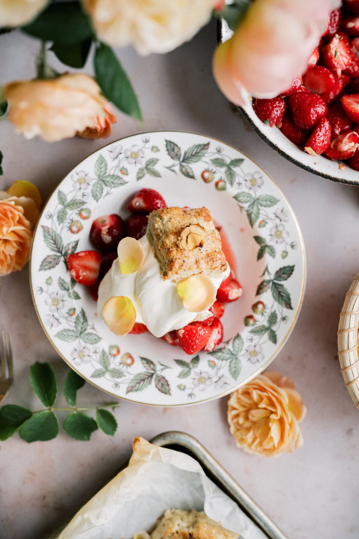 A floral-patterned plate holds a strawberry shortcake with biscuits, topped with whipped cream and fresh strawberries, garnished with flower petals. Yellow roses, a bowl of strawberries, and extra biscuits decorate the surrounding tray.