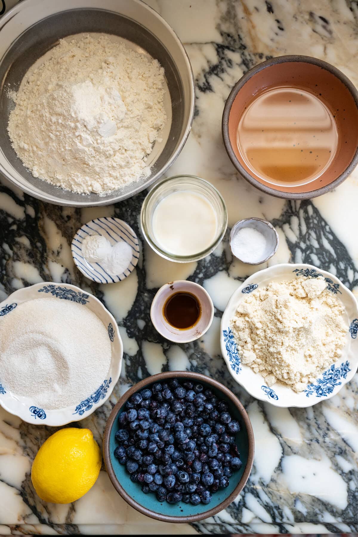 Ingredients for homemade blueberry crumb cake arranged on a marble countertop, including blueberries, lemon, flour, sugar, and butter.