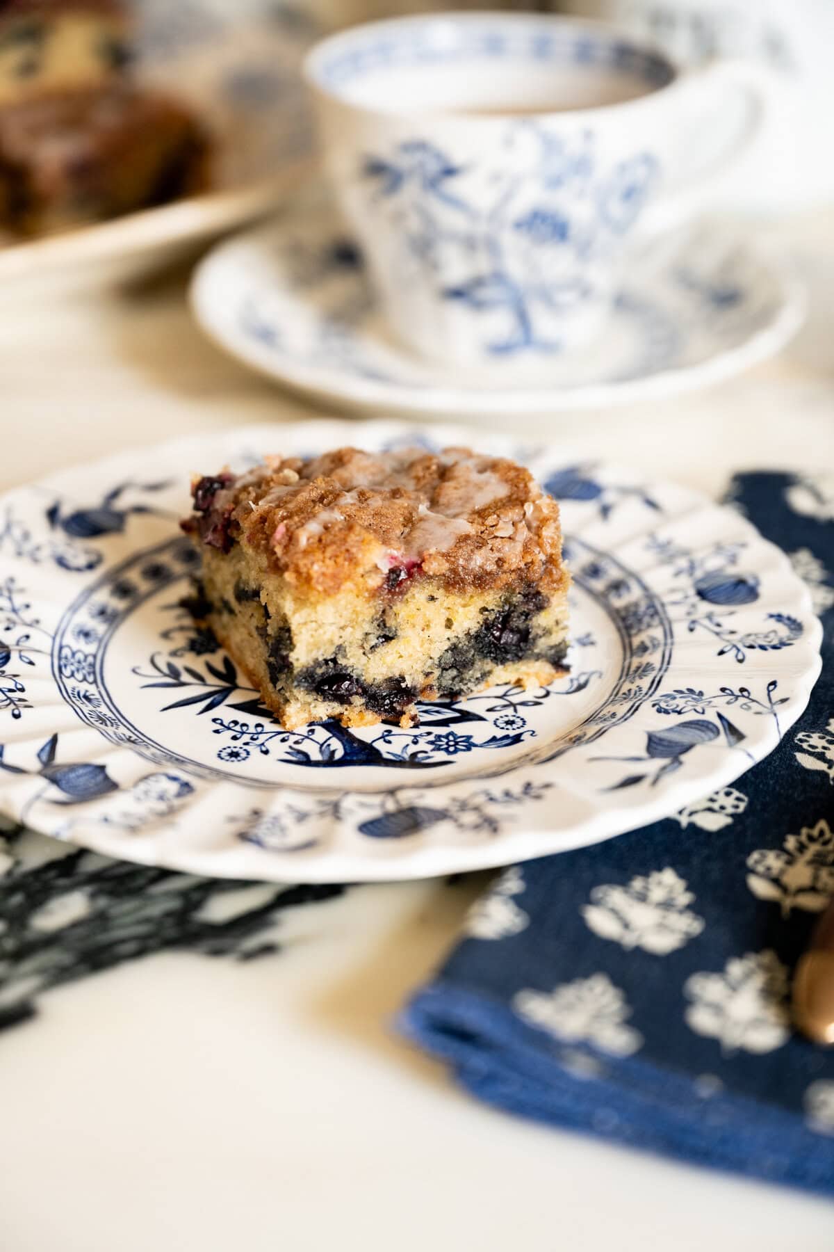 A slice of blueberry crumb coffee cake served on a blue-and-white floral plate with a cup of tea in the background.
