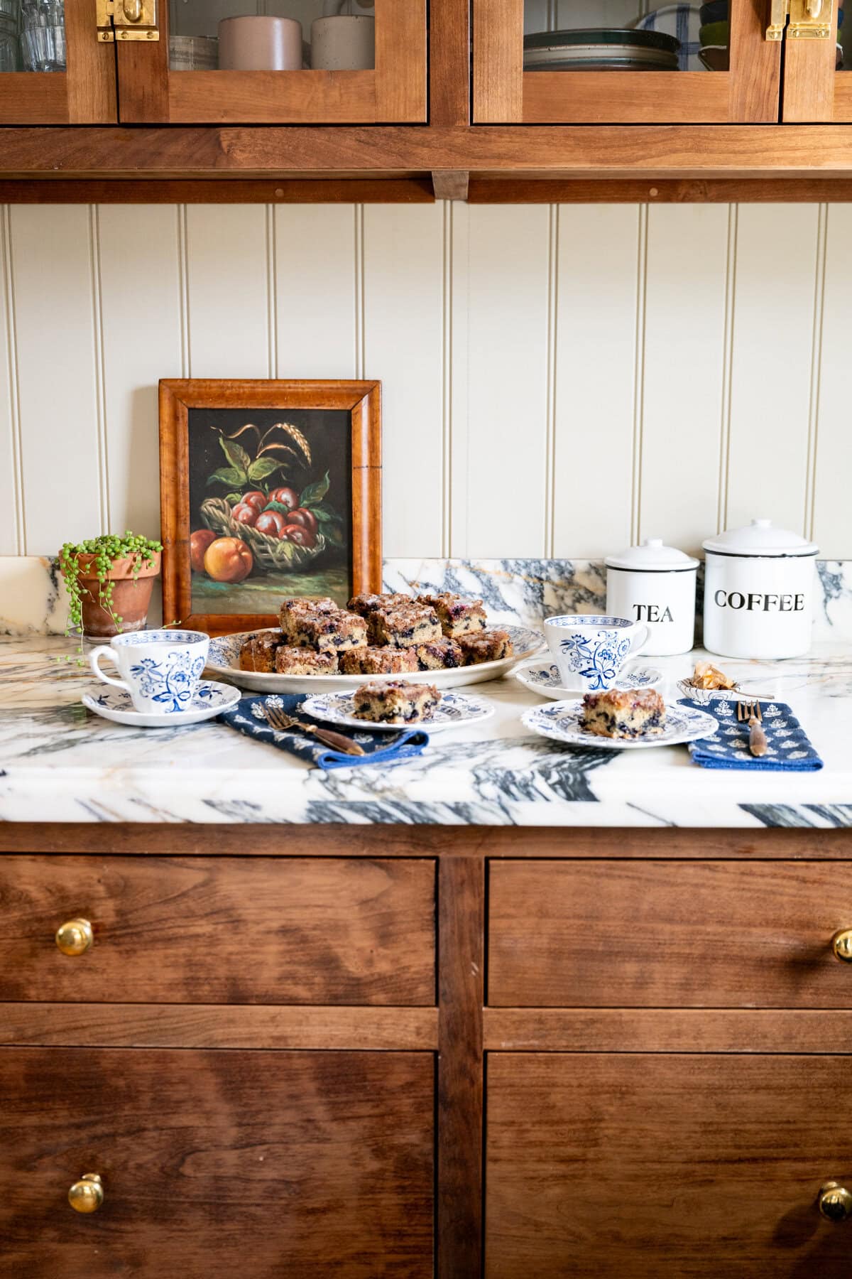 Homemade blueberry crumb cake arranged on a platter with individual servings, teacups, and vintage kitchen decor.