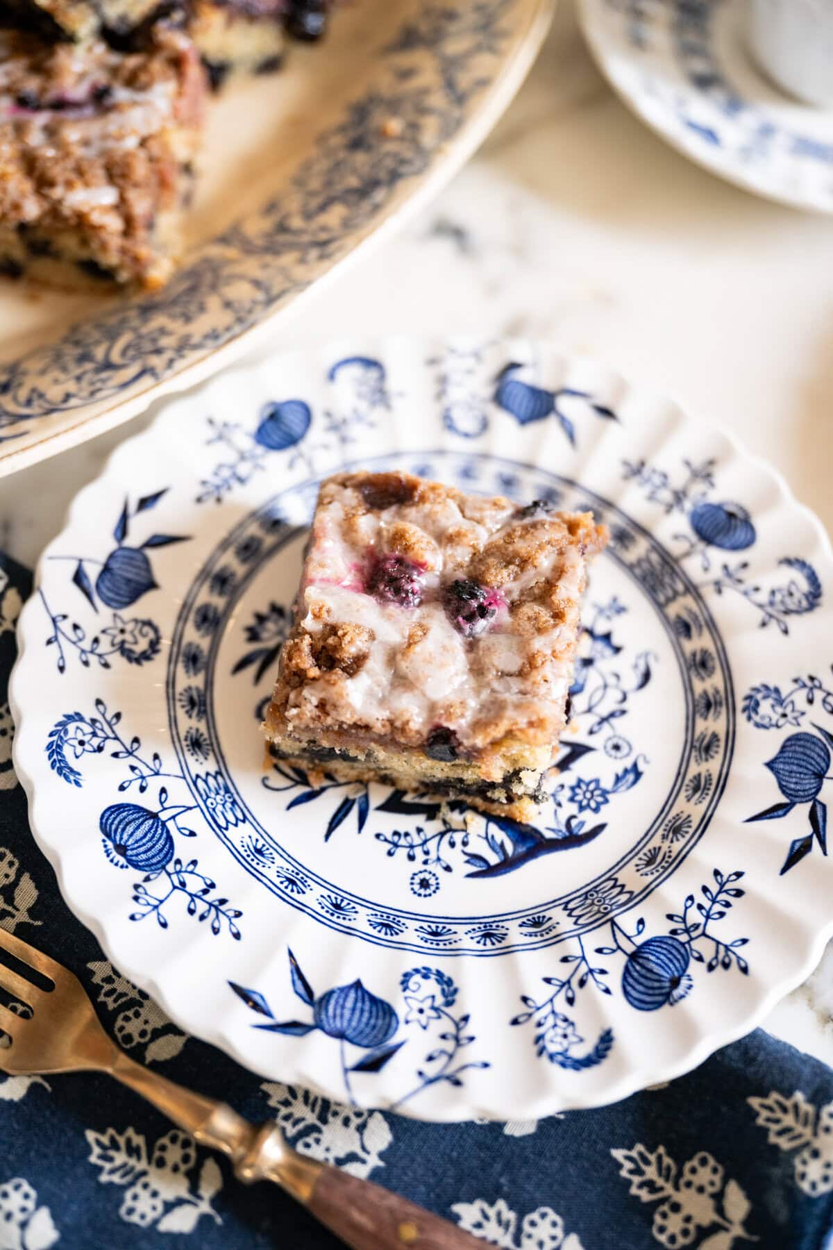 A square of blueberry crumb cake with lemon glaze served on a vintage blue-and-white patterned dessert plate.
