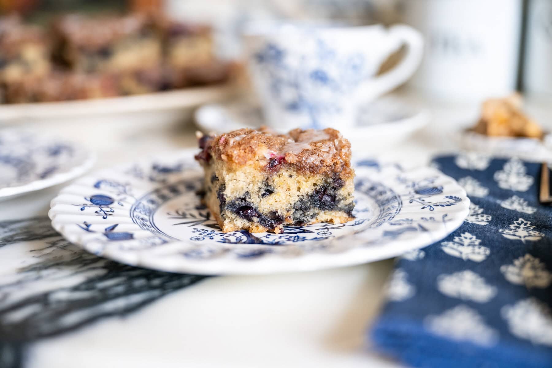 A piece of blueberry crumb cake with cinnamon streusel and lemon icing served on a floral dessert plate.